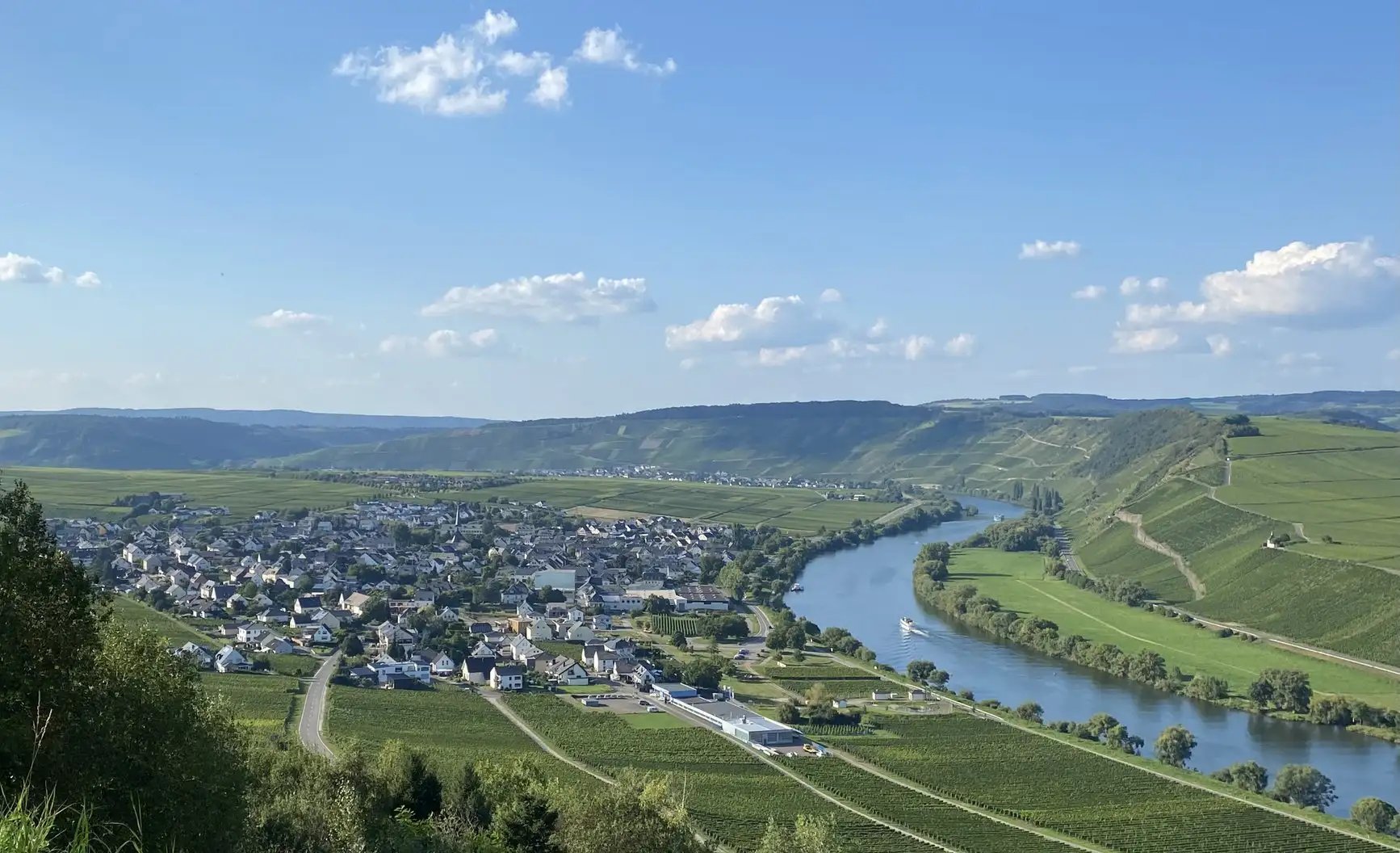 Ein Panoramablick auf einen Fluss, der sich durch grüne Hügel schlängelt, und eine kleine Stadt unter strahlend blauem Himmel.